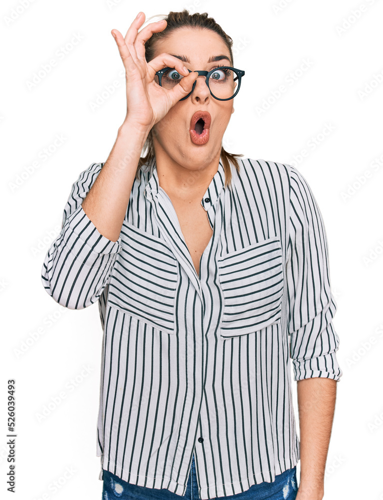 Young caucasian woman wearing business shirt and glasses doing ok gesture shocked with surprised face, eye looking through fingers. unbelieving expression.