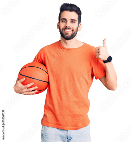 Young handsome man with beard hoilding basketball ball smiling happy and positive, thumb up doing excellent and approval sign photo