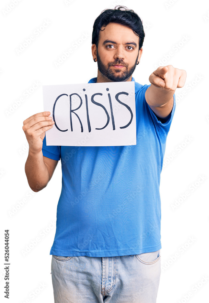 Young hispanic man holding crisis message paper pointing with finger to the camera and to you, confident gesture looking serious Young hispanic man holding crisis message paper pointing with finger to the camera and to you, confident gesture looking serious