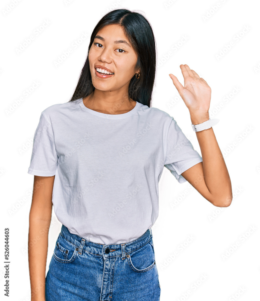 Beautiful young asian woman wearing casual white t shirt waiving saying hello happy and smiling, friendly welcome gesture