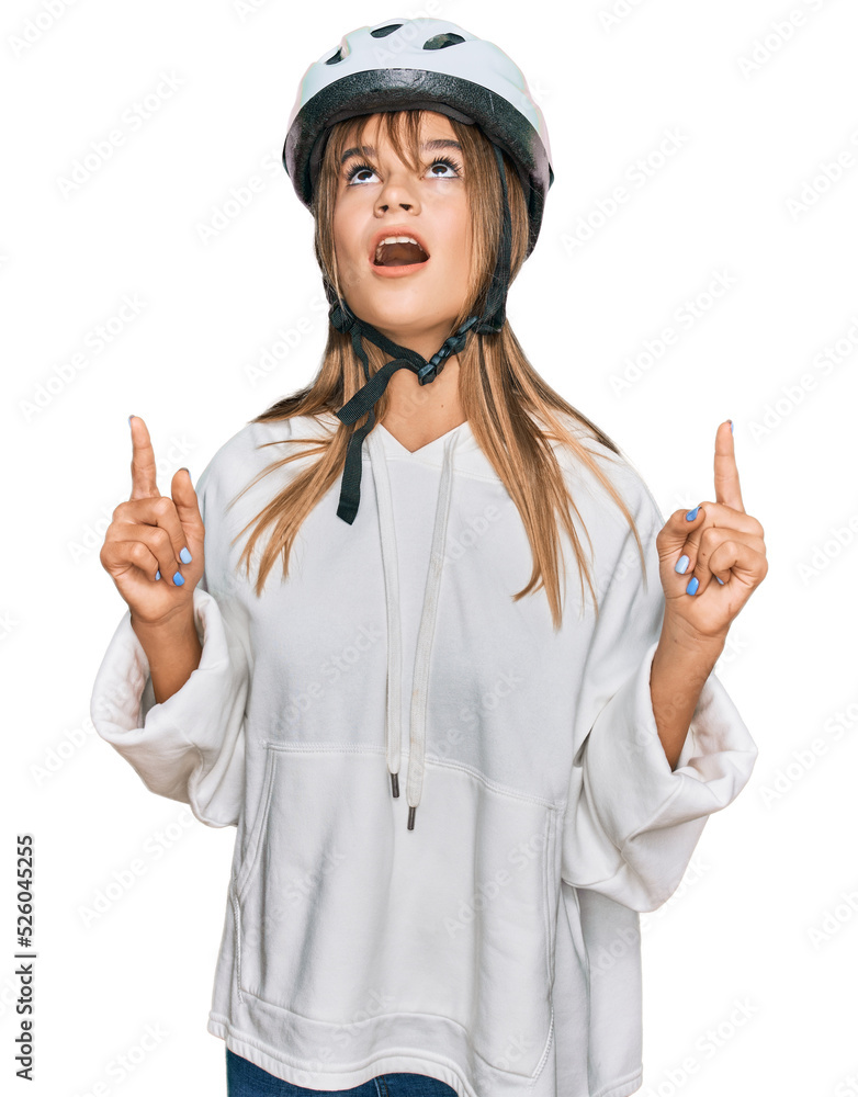 Teenager caucasian girl wearing bike helmet amazed and surprised looking up and pointing with fingers and raised arms. Teenager caucasian girl wearing bike helmet amazed and surprised looking up and pointing with fingers and raised arms.