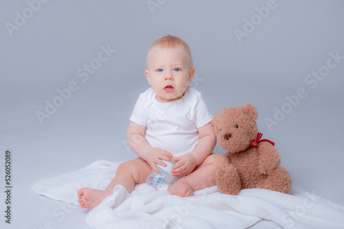 a baby boy in a white bodysuit sits with a teddy bear on a white background, holding a bottle of water , drinking water