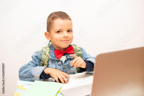 Little smiling schoolboy with a red bow tie and denim jacket sits at a table with a laptop.