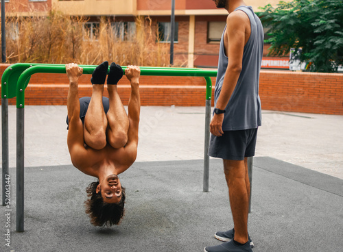 Two caucasian friends exercising in outdoor calisthenics bar doing upside down exercise.