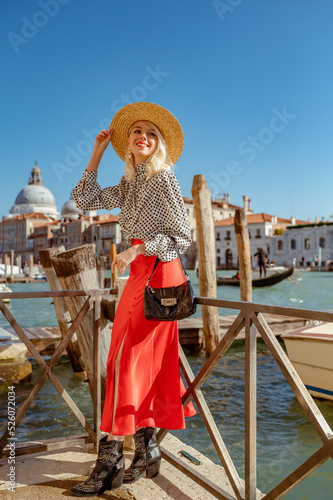 Happy smiling fashionable woman wearing straw hat, polka dot blouse, red skirt, ankle boots, with bag, posing in street of Venice. Fashion, travel, lifestyle conception. Full-length outdoor portrait