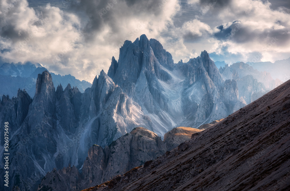 Epic rocky mountains at sunset in autumn. Mountain landscape with alpine mountains, orange grass on the hill, dramatic cloudy sky in fall. Aerial view of high rocks. Tre Cimein Dolomites, Italy