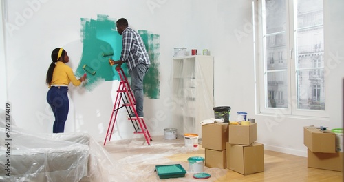 Fotografie Rear of African American young married couple wife and husband painting wall in green color using paint roller brush in new house with boxes