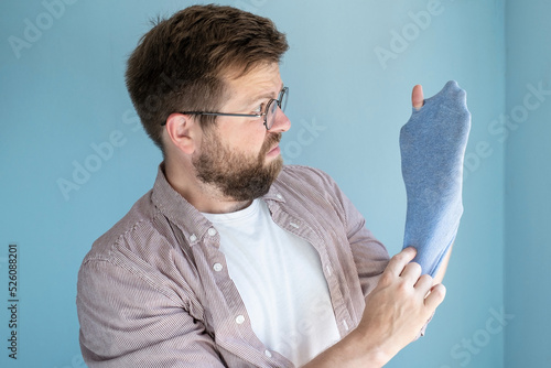 Frowning man put his holey worn sock on hand from which fingers stick out and looks with displeasure.