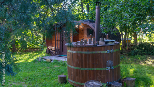 Outdoor wooden barrel sauna in the garden. In foreground wooden bathtub with fireplace. Wooden hot tub with spa in backyard.