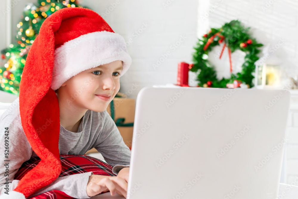 Cute boy in a Santa hat is typing on a laptop keyboard while lying in front of the Christmas tree at home. Smiling child looking at screen and chatting with friends during the winter holidays.