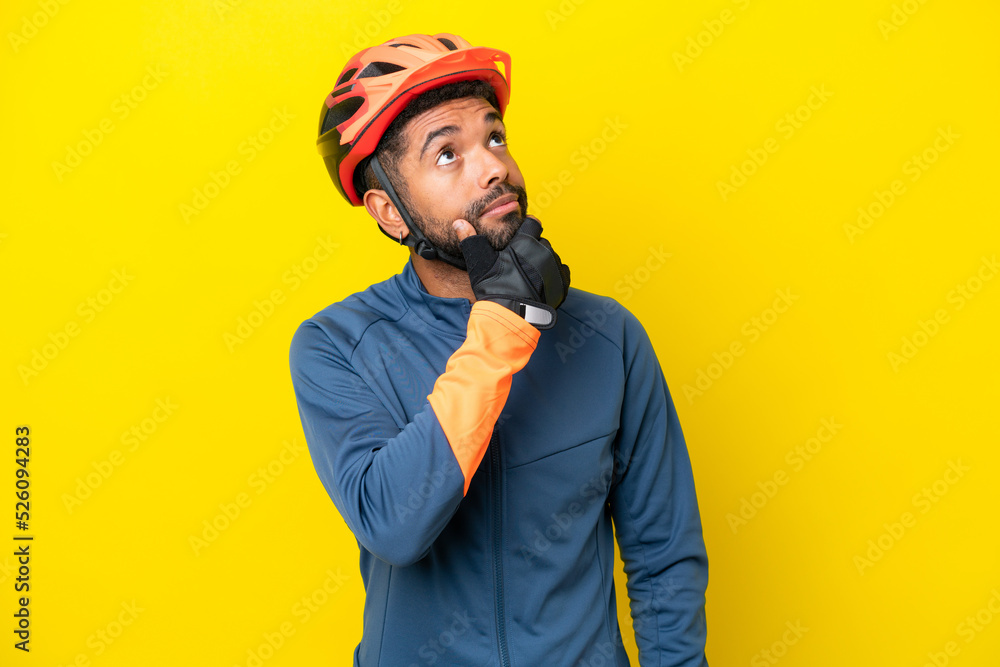 Young cyclist Brazilian man isolated on yellow background having doubts Young cyclist Brazilian man isolated on yellow background having doubts
