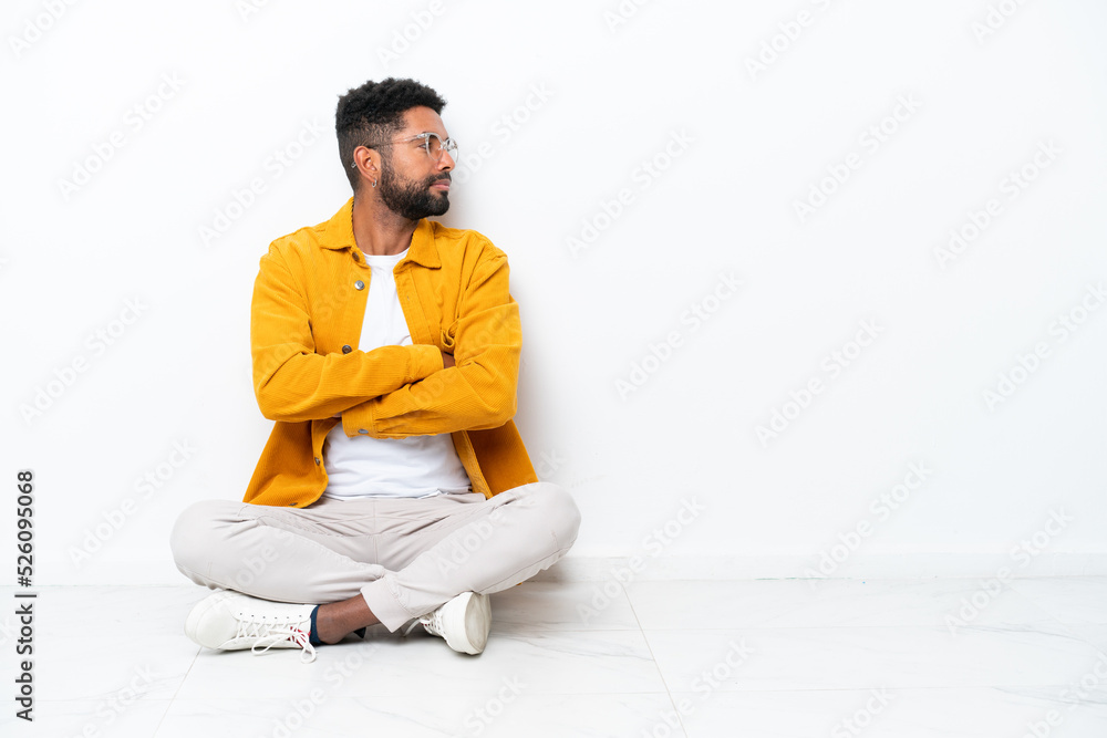 Young Brazilian man sitting on the floor isolated on white background in lateral position Young Brazilian man sitting on the floor isolated on white background in lateral position