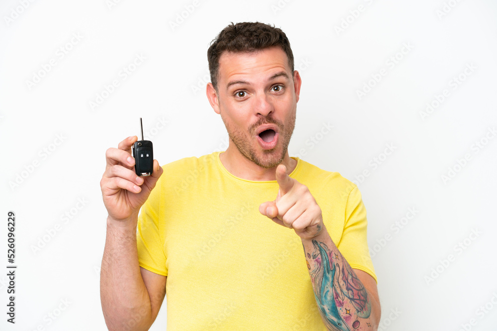 Young man holding car keys isolated on white background surprised and pointing front Young man holding car keys isolated on white background surprised and pointing front