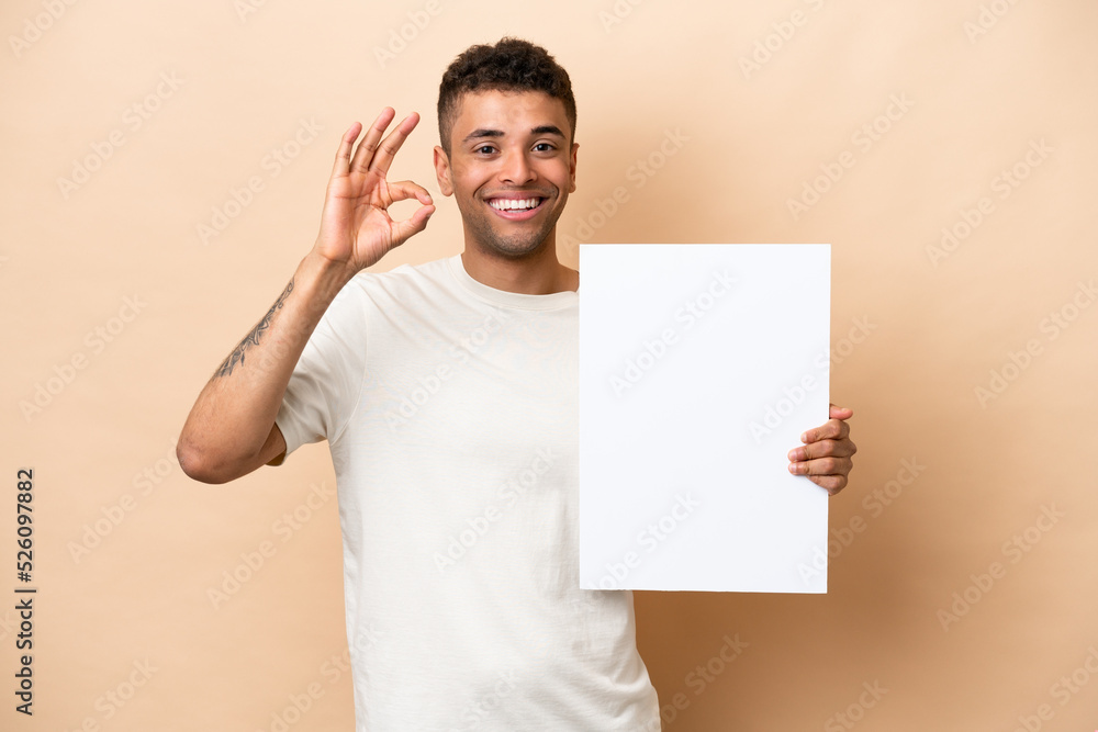 Young Brazilian man isolated on beige background holding an empty placard and doing OK sign Young Brazilian man isolated on beige background holding an empty placard and doing OK sign