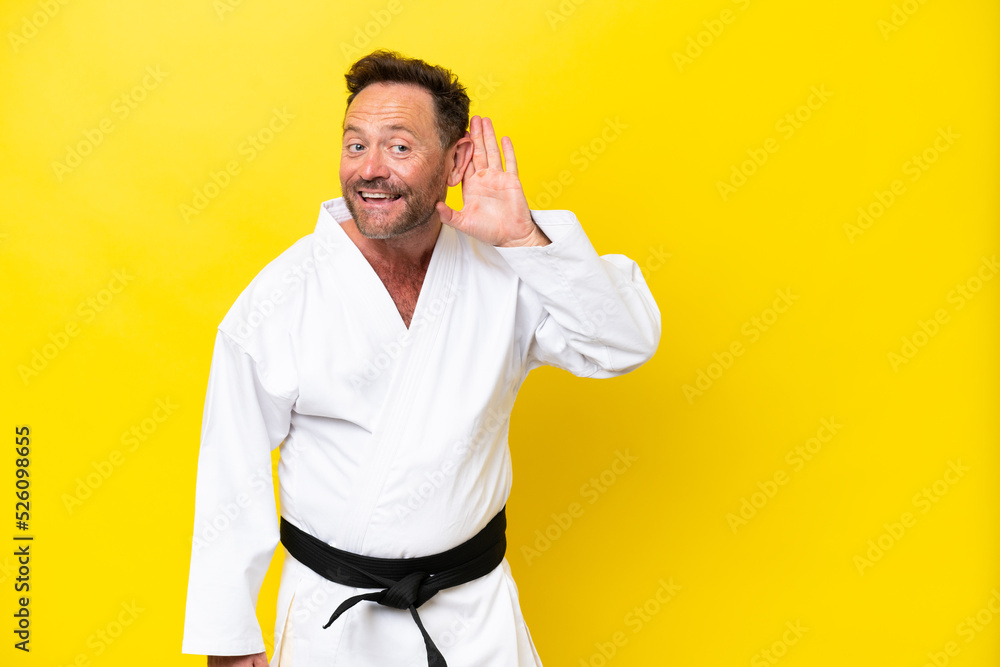 Middle age caucasian man doing karate isolated on yellow background listening to something by putting hand on the ear Middle age caucasian man doing karate isolated on yellow background listening to something by putting hand on the ear