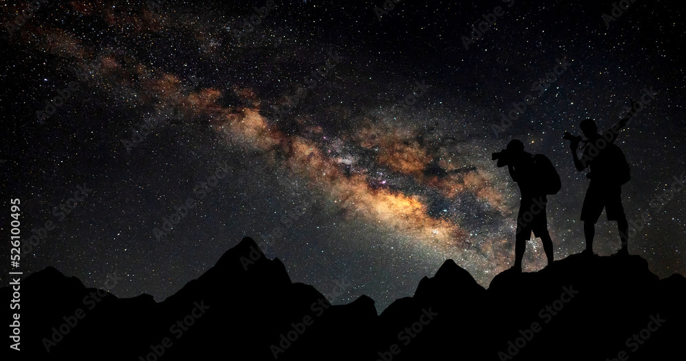 Silhouette of Two Cameraman standing on rock.Warm Color night sky with stars and milky way with man on the mountain. High Rocks.Background with galaxy and silhouette of a man. Many stars.