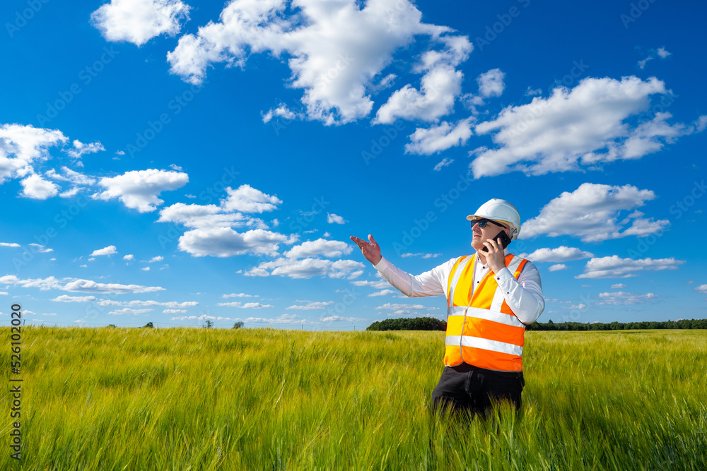 Builder is talking on phone. Man in builder's uniform. Human stands among tall grass. Man inspects area in front of construction. Selection land for construction. Construction worker under blue sky Builder is talking on phone. Man in builder's uniform. Human stands among tall grass. Man inspects area in front of construction. Selection land for construction. Construction worker under blue sky
