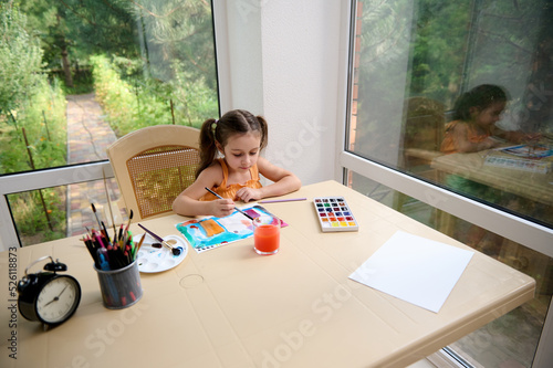 Adorable child, elementary age girl sitting at a table and painting picture with watercolors and paintbrush. Kids education and development. Drawing. Art classes and courses for telented children photo