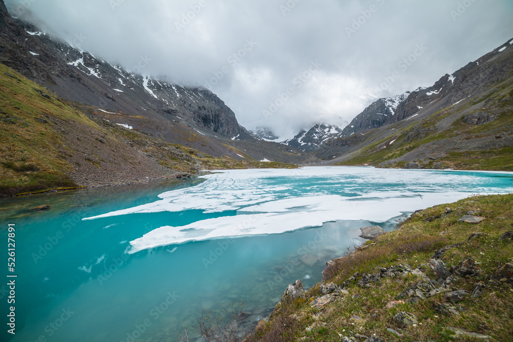 Atmospheric mountain landscape with frozen alpine lake and high snowy mountains. Awesome overcast scenery with icy mountain lake on background of snow mountains in low clouds. Scenic view to ice lake.
