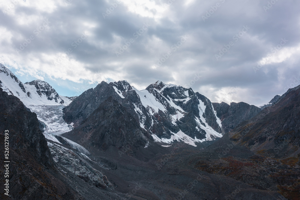 Atmospheric landscape with large snow mountain range with glacier and icefall in dramatic cloudy sky. Awesome high snowy mountains under rainy clouds. White snow on black rocks in overcast weather.