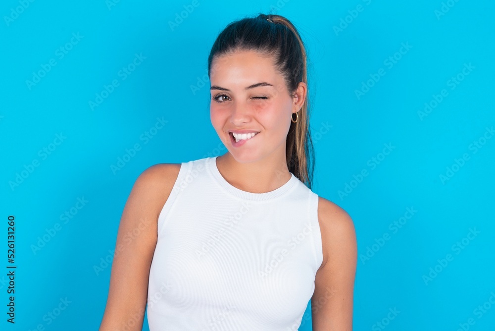 Fototapeta premium Amazed beautiful brunette woman wearing white tank top over blue background bitting lip and looking tricky to empty space.