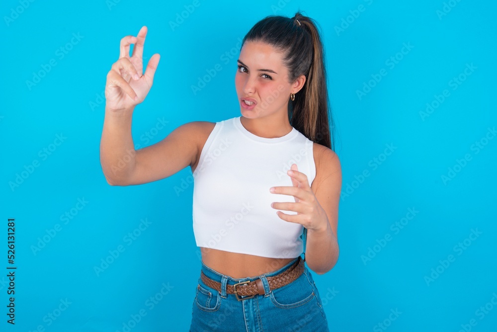 beautiful brunette woman wearing white tank top over blue background Shouting frustrated with rage, hands trying to strangle, yelling mad. beautiful brunette woman wearing white tank top over blue background Shouting frustrated with rage, hands trying to strangle, yelling mad.