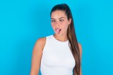 beautiful brunette woman wearing white tank top over blue background showing grimace face crossing eyes and showing tongue. Being funny and crazy