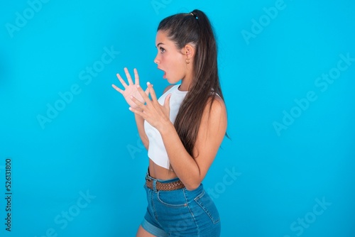 beautiful brunette woman wearing white tank top over blue background shouts loud, keeps eyes opened and hands tense.