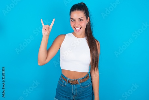 beautiful brunette woman wearing white tank top over blue background doing a rock gesture and smiling to the camera. Ready to go to her favorite band concert.
