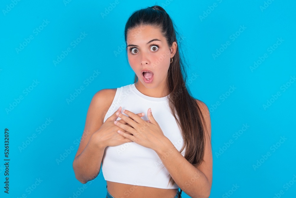 Scared beautiful brunette woman wearing white tank top over blue looks with frightened expression, keeps hands on chest, being puzzled to notice something strange, People, hush reaction and emotions. Scared beautiful brunette woman wearing white tank top over blue looks with frightened expression, keeps hands on chest, being puzzled to notice something strange, People, hush reaction and emotions.