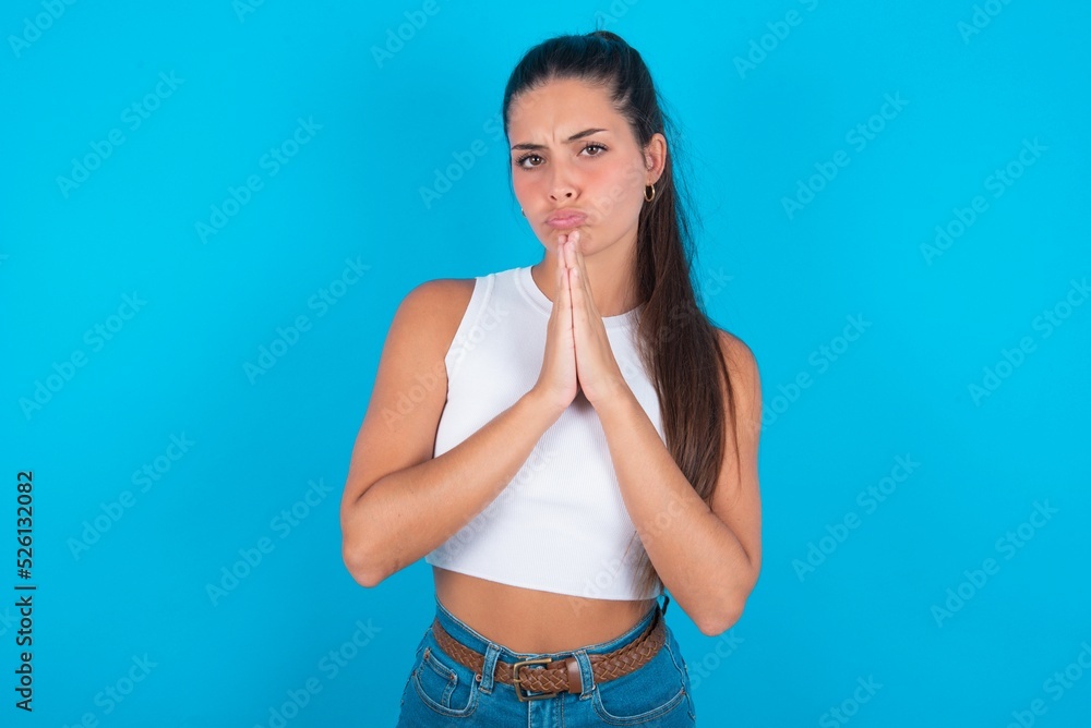 Positive beautiful brunette woman wearing white tank top over blue background smiles happily, glad to receive pleasant news from interlocutor, keeps palms together, People emotions concept. Positive beautiful brunette woman wearing white tank top over blue background smiles happily, glad to receive pleasant news from interlocutor, keeps palms together, People emotions concept.
