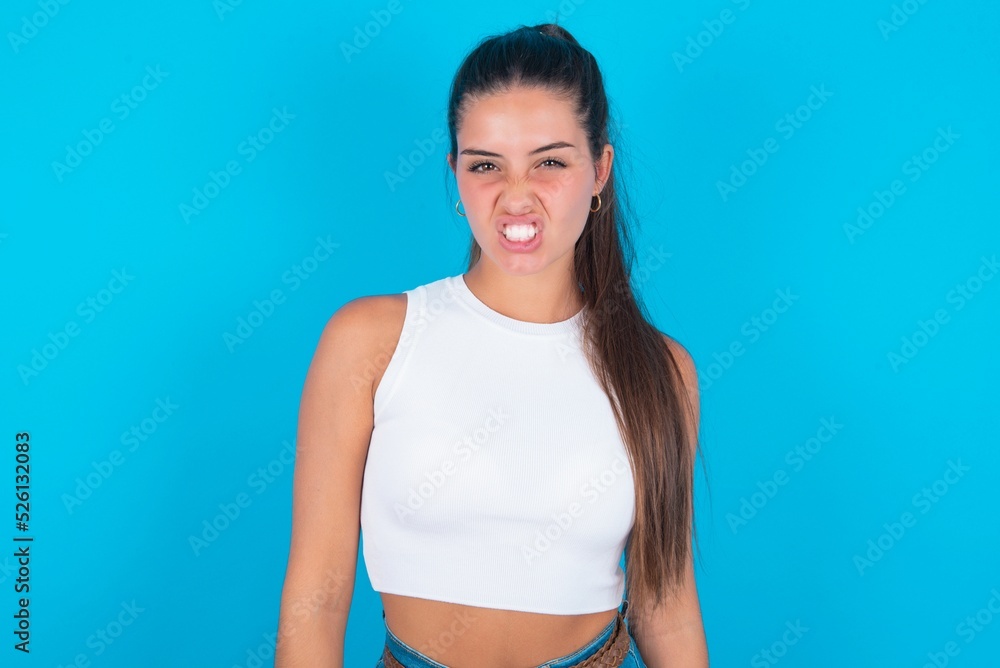 beautiful brunette woman wearing white tank top over blue background keeps teeth clenched, frowns face in dissatisfaction, irritated because of much duties. beautiful brunette woman wearing white tank top over blue background keeps teeth clenched, frowns face in dissatisfaction, irritated because of much duties.
