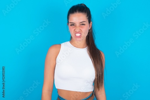 beautiful brunette woman wearing white tank top over blue background keeps teeth clenched, frowns face in dissatisfaction, irritated because of much duties. © Roquillo