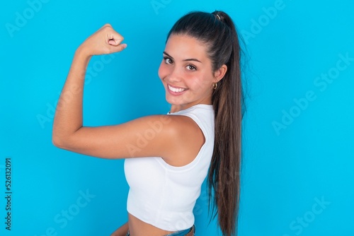 Portrait of powerful cheerful beautiful brunette woman wearing white tank top over blue background showing muscles.