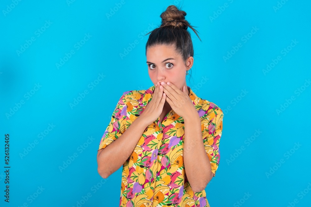 beautiful brunette woman wearing colourful shirt over blue background keeps hands on mouth, looks with eyes full of disbelief, being puzzled with amount of work beautiful brunette woman wearing colourful shirt over blue background keeps hands on mouth, looks with eyes full of disbelief, being puzzled with amount of work