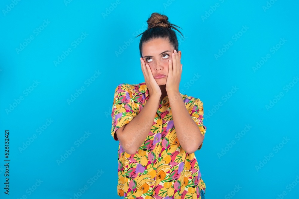 beautiful brunette woman wearing colourful shirt over blue background keeps hands on cheeks has bored displeased expression. Stressed hopeless model beautiful brunette woman wearing colourful shirt over blue background keeps hands on cheeks has bored displeased expression. Stressed hopeless model
