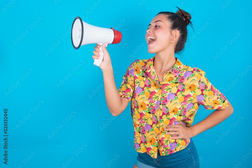 Funny beautiful brunette woman wearing colourful shirt over blue background People sincere emotions lifestyle concept. Mock up copy space. Screaming in megaphone. Funny beautiful brunette woman wearing colourful shirt over blue background People sincere emotions lifestyle concept. Mock up copy space. Screaming in megaphone.