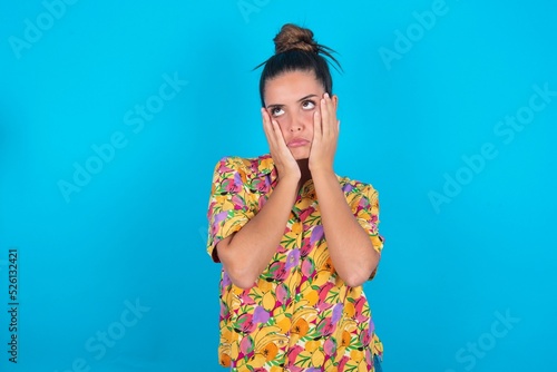 beautiful brunette woman wearing colourful shirt over blue background keeps hands on cheeks has bored displeased expression. Stressed hopeless model
