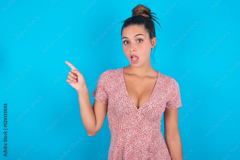 Emotive beautiful brunette woman wearing floral dress over blue wall keeps jaw dropped from shock demonstrates amazing promo points right on blank space demonstrates big shopping sale. Advertisement Emotive beautiful brunette woman wearing floral dress over blue wall keeps jaw dropped from shock demonstrates amazing promo points right on blank space demonstrates big shopping sale. Advertisement