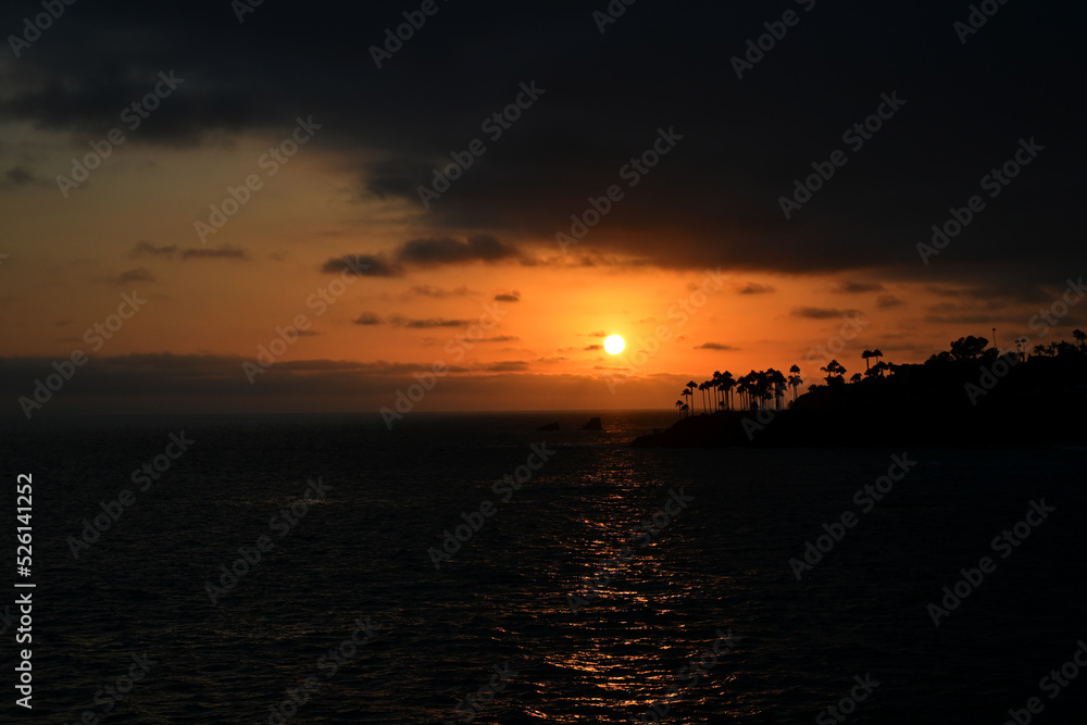 A bright orange sky as the sun sets between layers of clouds over Twin Points, Laguna Beach, California.