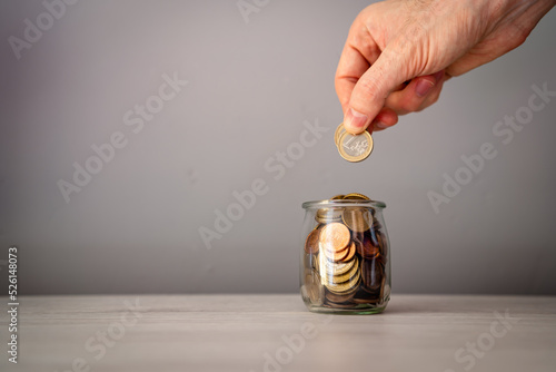 Photos Man's hand adding coins inside a glass jar on a light wooden table, to save or donate