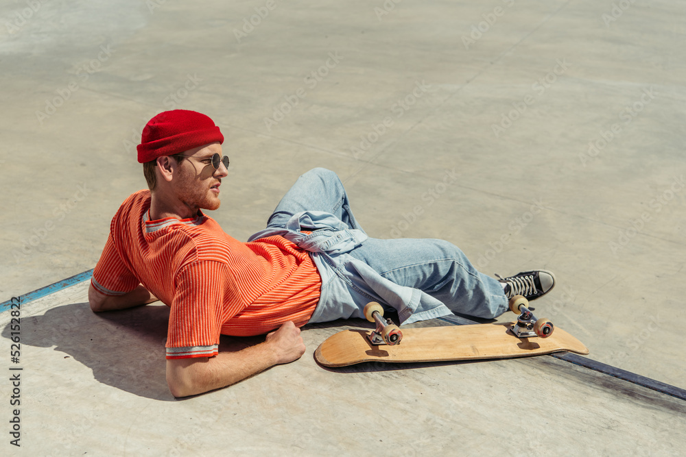 young man in orange t-shirt and beanie resting near skateboard in park young man in orange t-shirt and beanie resting near skateboard in park