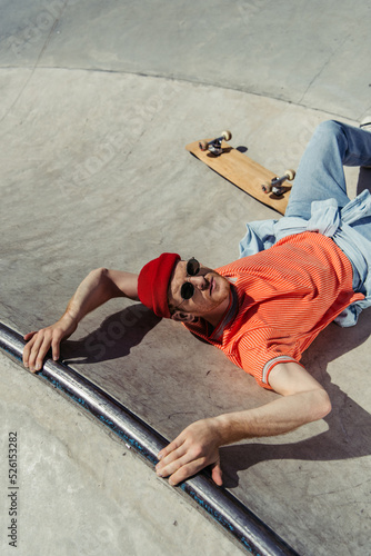 high angle view of man in sunglasses and beanie looking at camera while lying on ramp near skate
