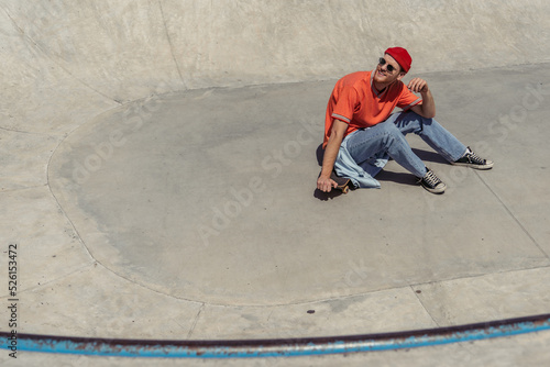 high angle view of man in red beanie and sunglasses sitting on skateboard in skate park