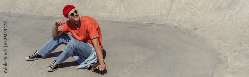 smiling man in fashionable clothes sitting on skateboard and looking away, banner © LIGHTFIELD STUDIOS