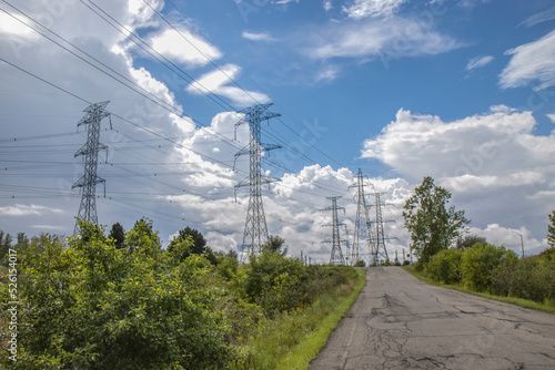 Rows of electrical high voltage transmission pylons and lines, paved road in foreground, daytime sunny with clouds, nobody