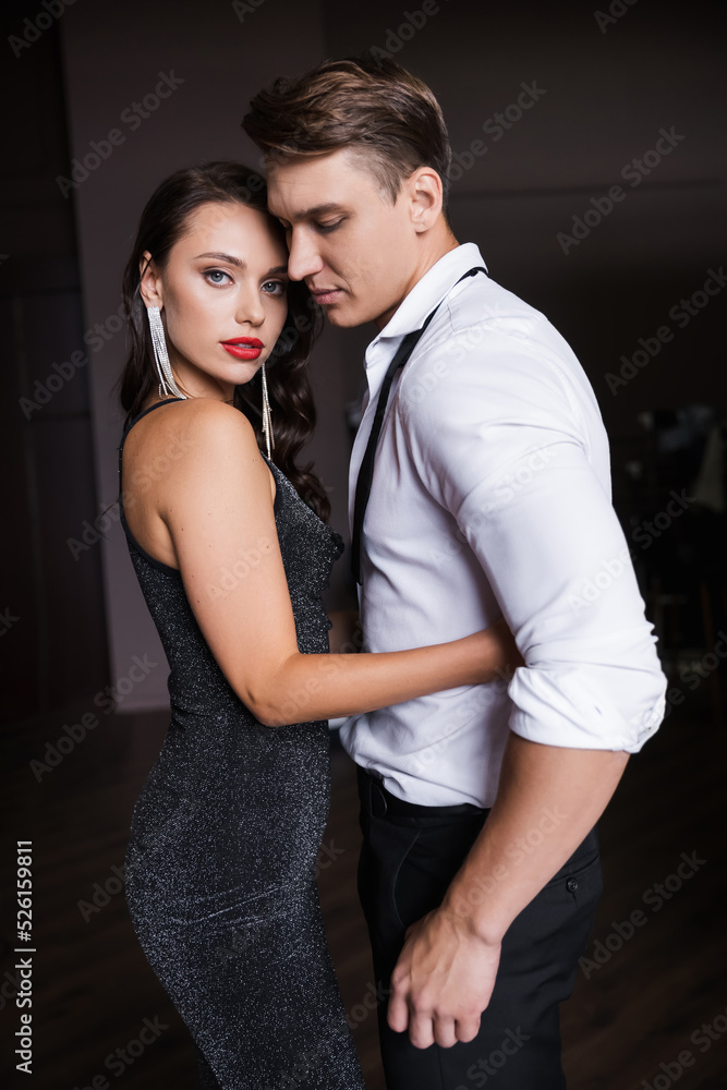 Seductive woman in dress hugging elegant boyfriend and looking at camera in evening