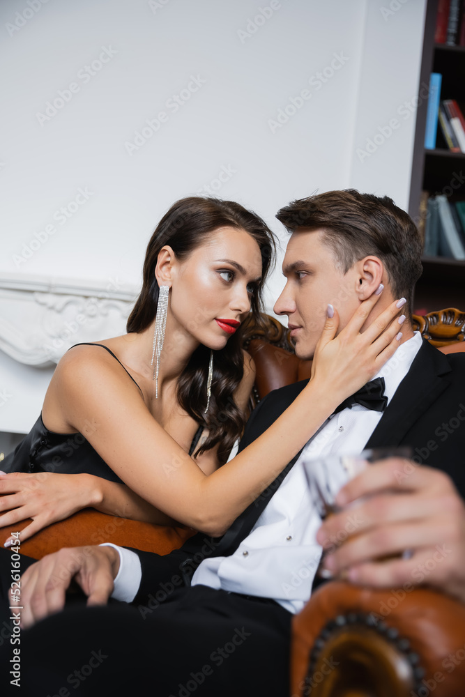Sensual woman in silk dress touching boyfriend in suit holding blurred glass at home Sensual woman in silk dress touching boyfriend in suit holding blurred glass at home