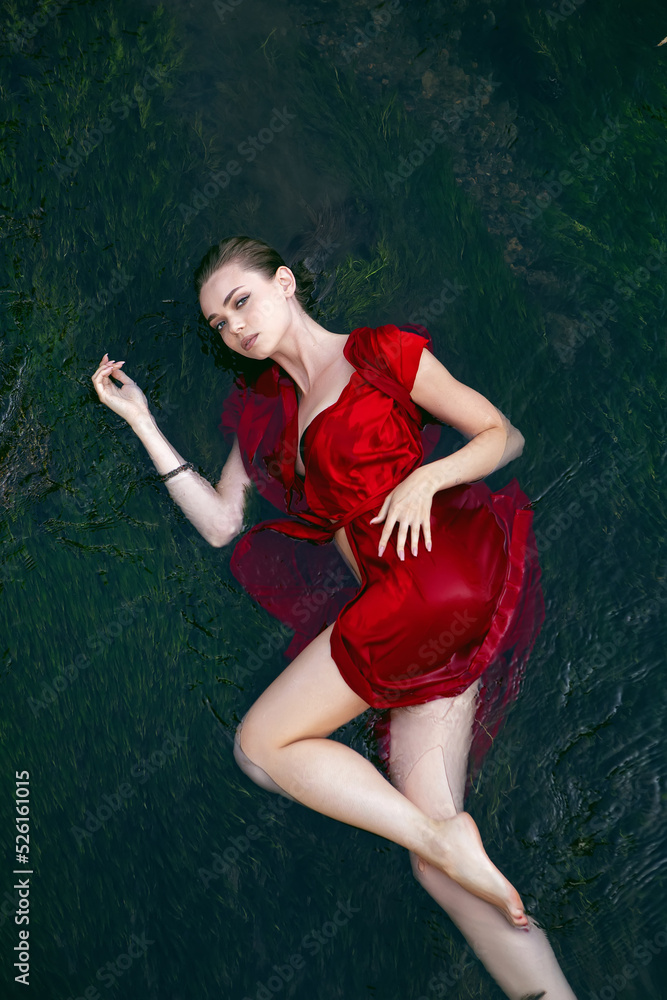 Art beautiful romantic portrait of a sexy young woman in a red dress lying in a river with green algae in summer in nature during the day.