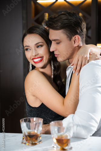 Cheerful woman with red lips hugging boyfriend near blurred glasses of whiskey in bar